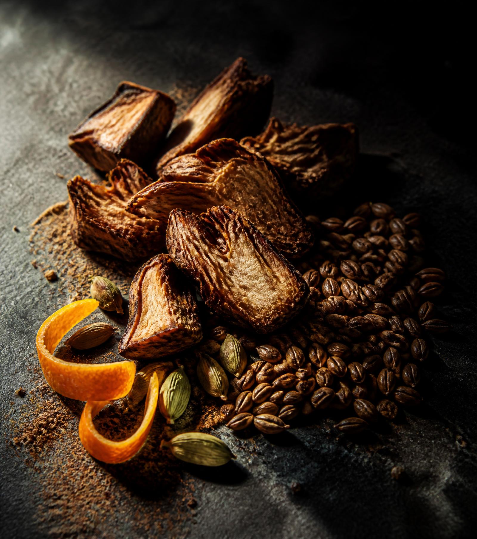 Roasted chicory root, barley, cardamom pods and orange peel on a dark stone slab under raking light